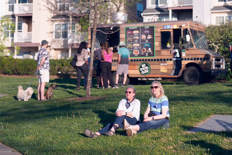 Attendees sit on grass enjoying music while other attendees wait in line for food truck in the background