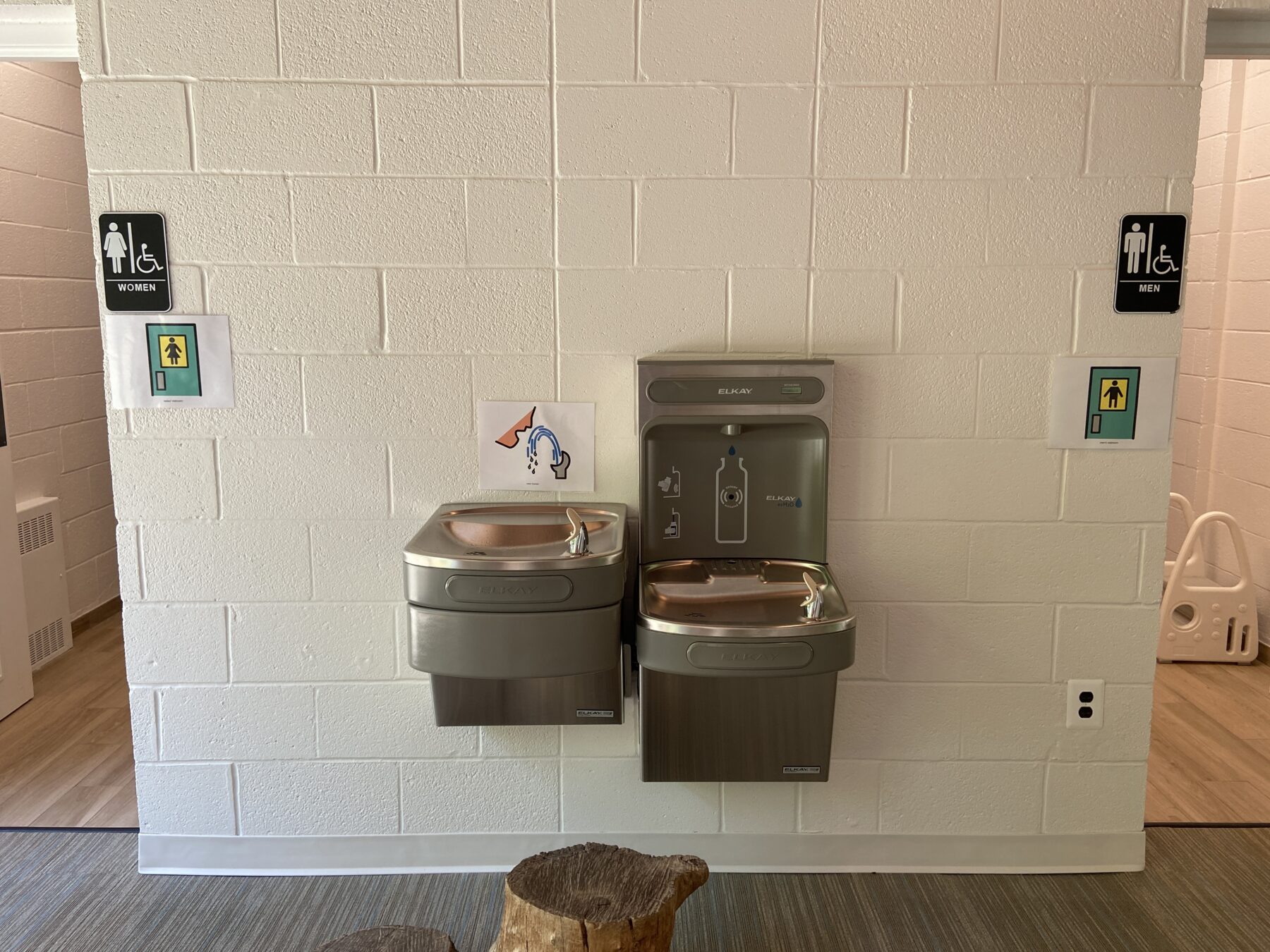 A picture of the outisde of the bathrooms with a water fountain in between the two