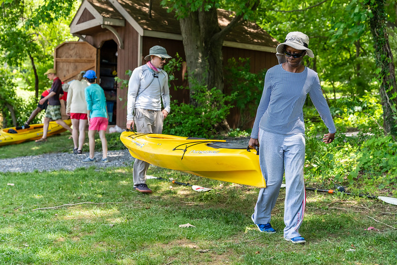 Two adults each work together to carry a yellow kayak. They are walking across a green lawn.