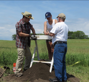 3 people surrounding a tray on top of a small pile of dirt, grassy field in the background