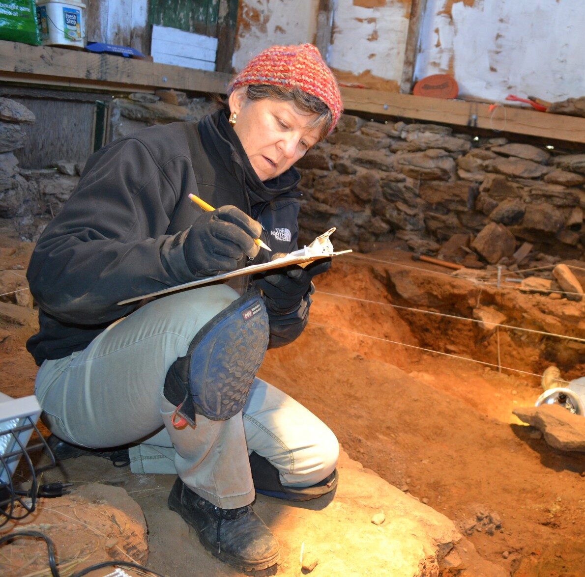 A woman sits in a dig site writing on a clipboard