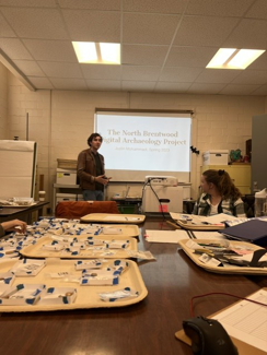 A man standing in front of a projector. A table with trays of archeology materials in the foreground