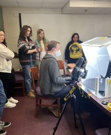 A group of students standing behind and observing a man in front of them who appears to be examining an object at a table