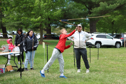 A boy standing with his arm raised using an atlatl while 4 people stand behind him and watch
