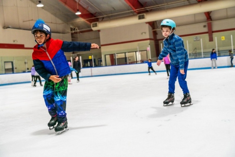 Two kids in the foreground ice skating with jackets and helmets on. More kids skating in the background