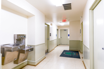 Interior view of Viers Mill PAB that is showing the hallway with the restroom. Displayed next to the doors are the "women's" "men's" and "stairwell" signs. There are two water fountains on the wall to the left