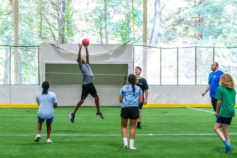 Six people playing a ball game on a sport field the person on the left is midair catching the ball
